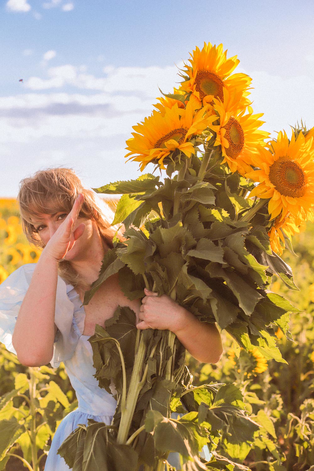A woman in a blue dress peeks out from behind a bunch of sunflowers